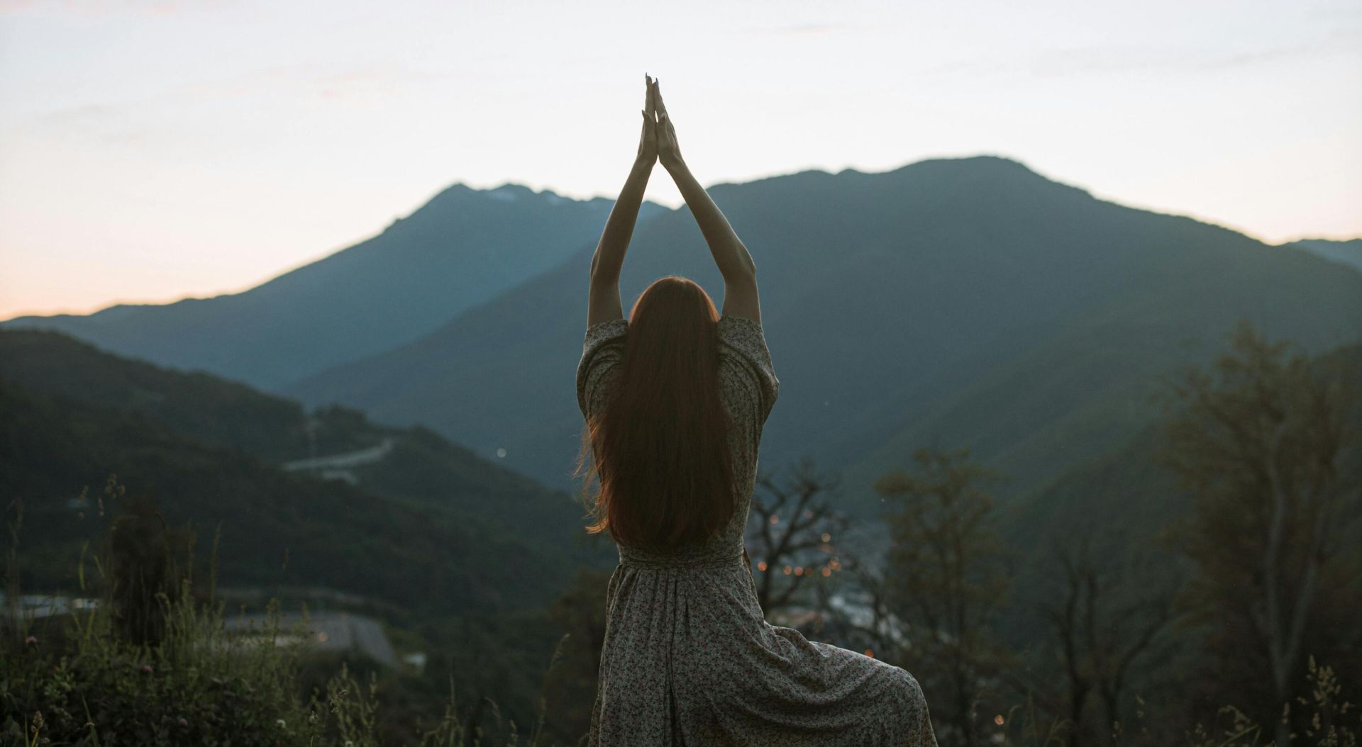 A woman in a dress performs tree pose against a mountainous sunrise backdrop, adding serenity and strength.